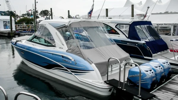 Bateau de luxe bleu et blanc équipé d’une toile grise sur mesure et d’un pare-brise transparent, amarré dans une marina avec moteurs Mercury en arrière-plan.