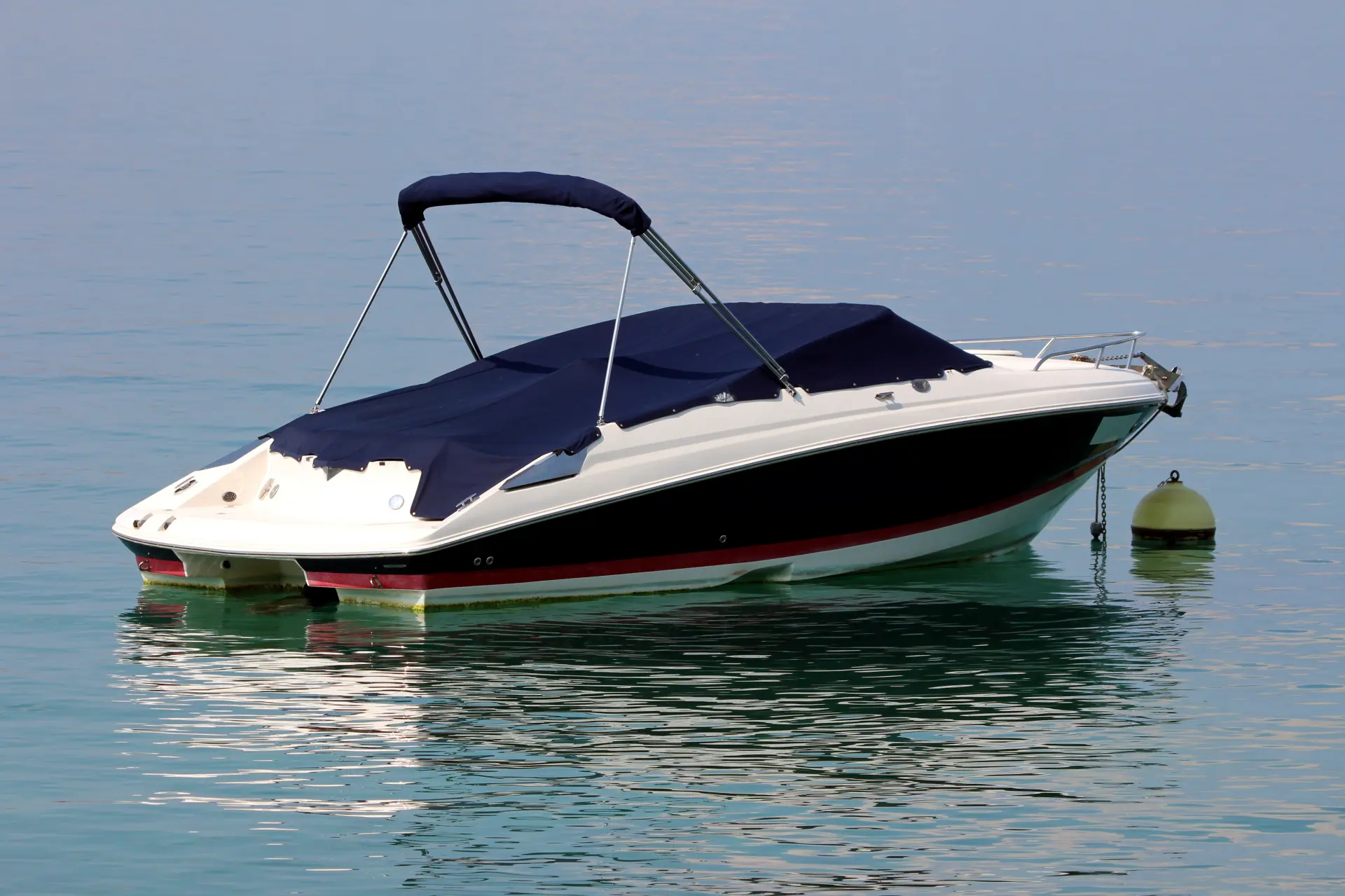 Petit bateau de plaisance blanc et noir équipé d’une toile de protection bleue foncée, amarré à une bouée sur une eau calme.