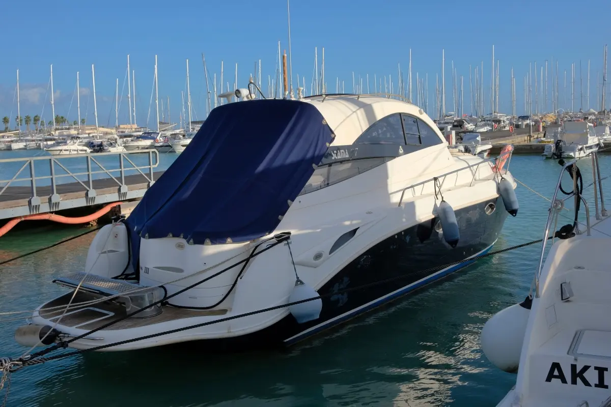 Yacht moderne de couleur blanche et bleue, amarré dans une marina, recouvert d’une toile de protection bleue tendue à l’arrière, entouré d’autres bateaux à quai.